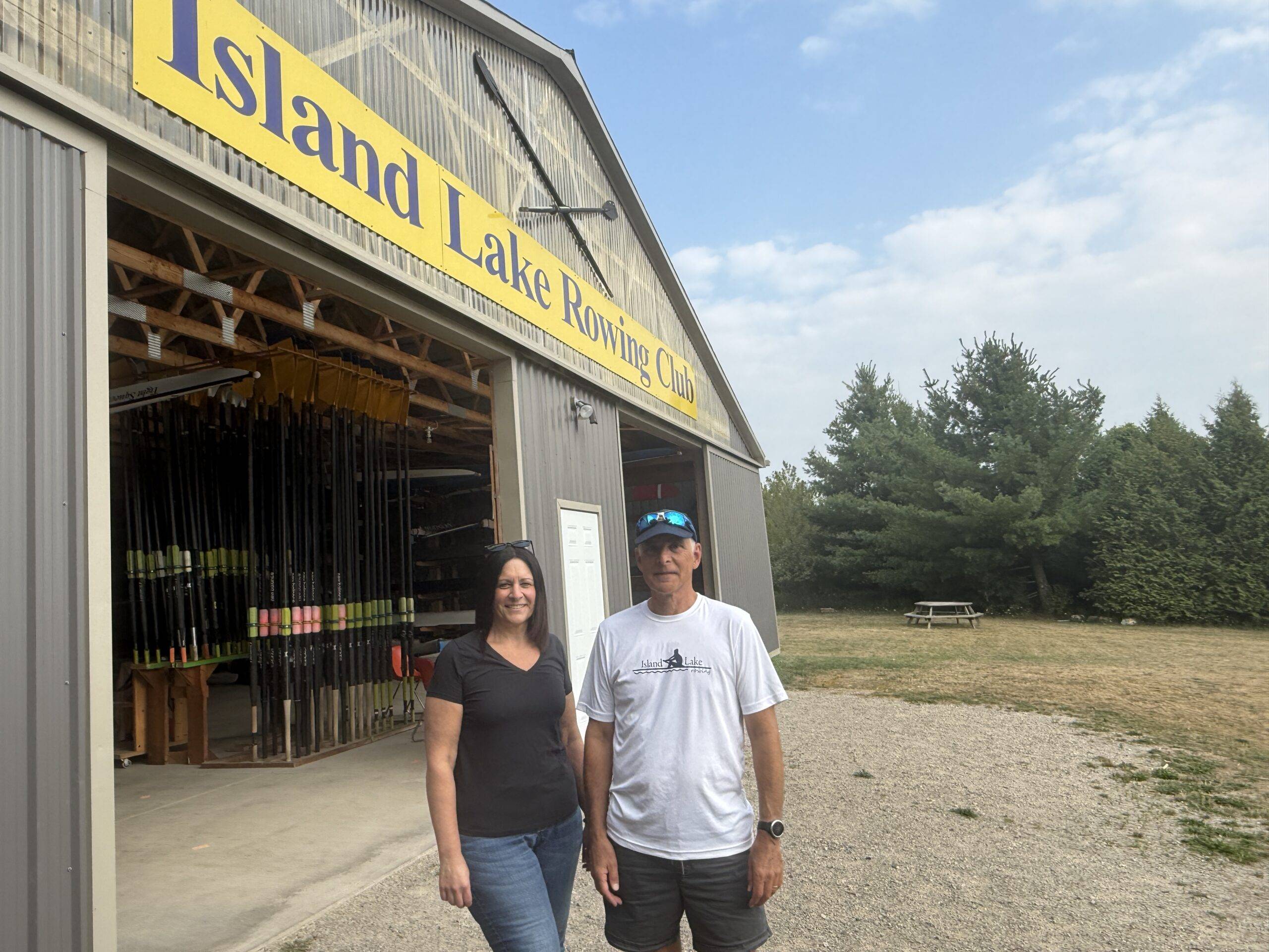 Two coaches stand in front of the Island Lake Rowing Club boathouse, with rowing equipment visible inside and trees in the background.