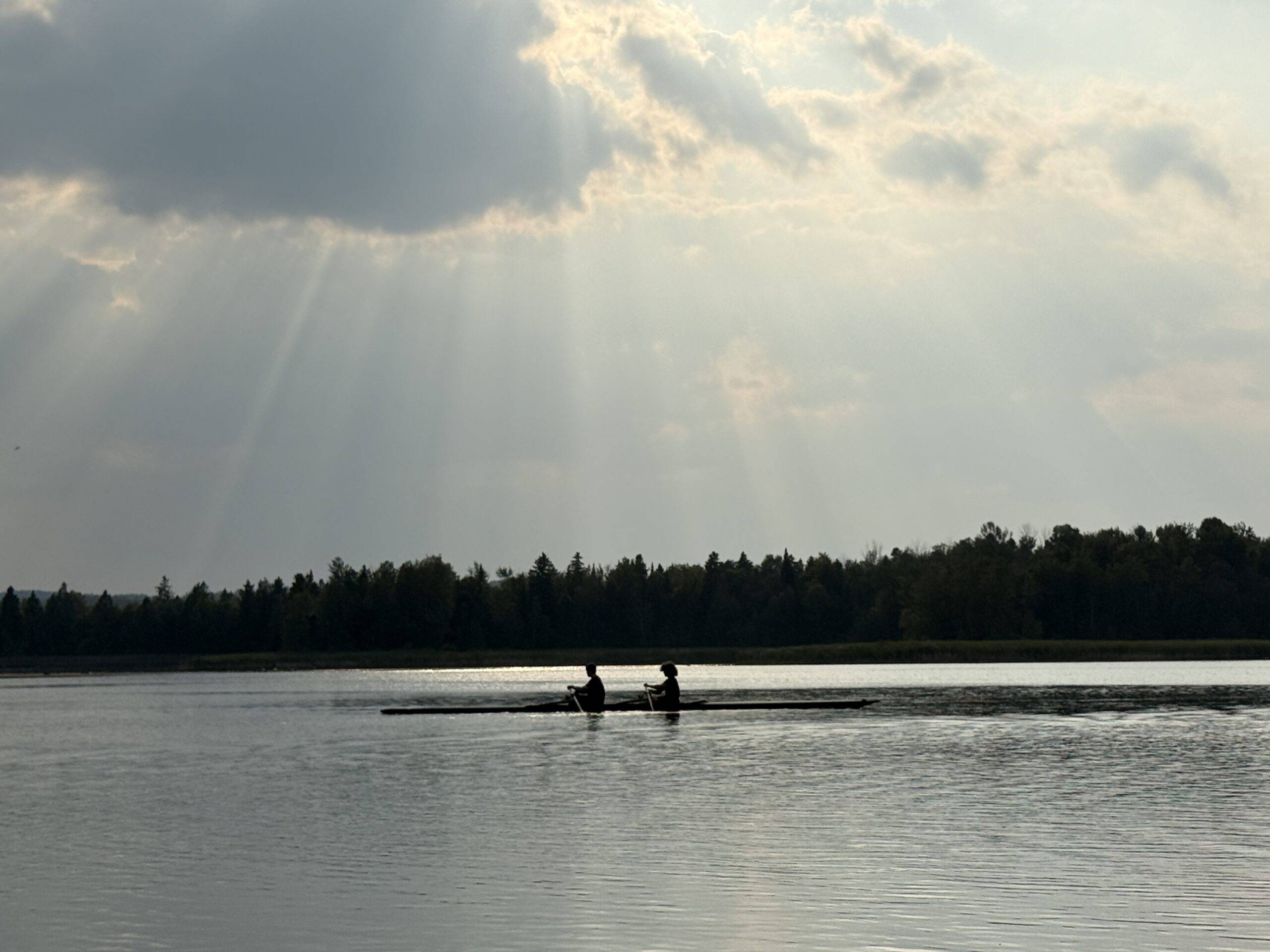 Two people row on a calm lake with a forested shoreline in the background and sun rays breaking through a partly cloudy sky.