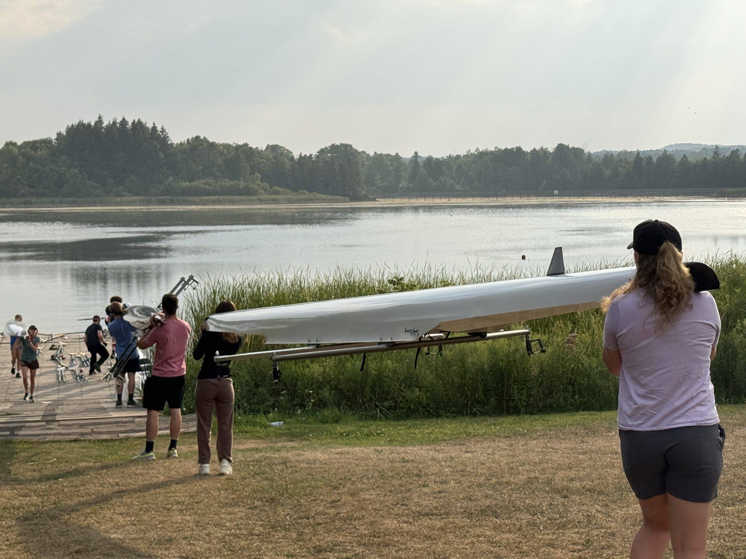 A group of people carry a long rowing boat toward the lake, surrounded by trees and greenery.