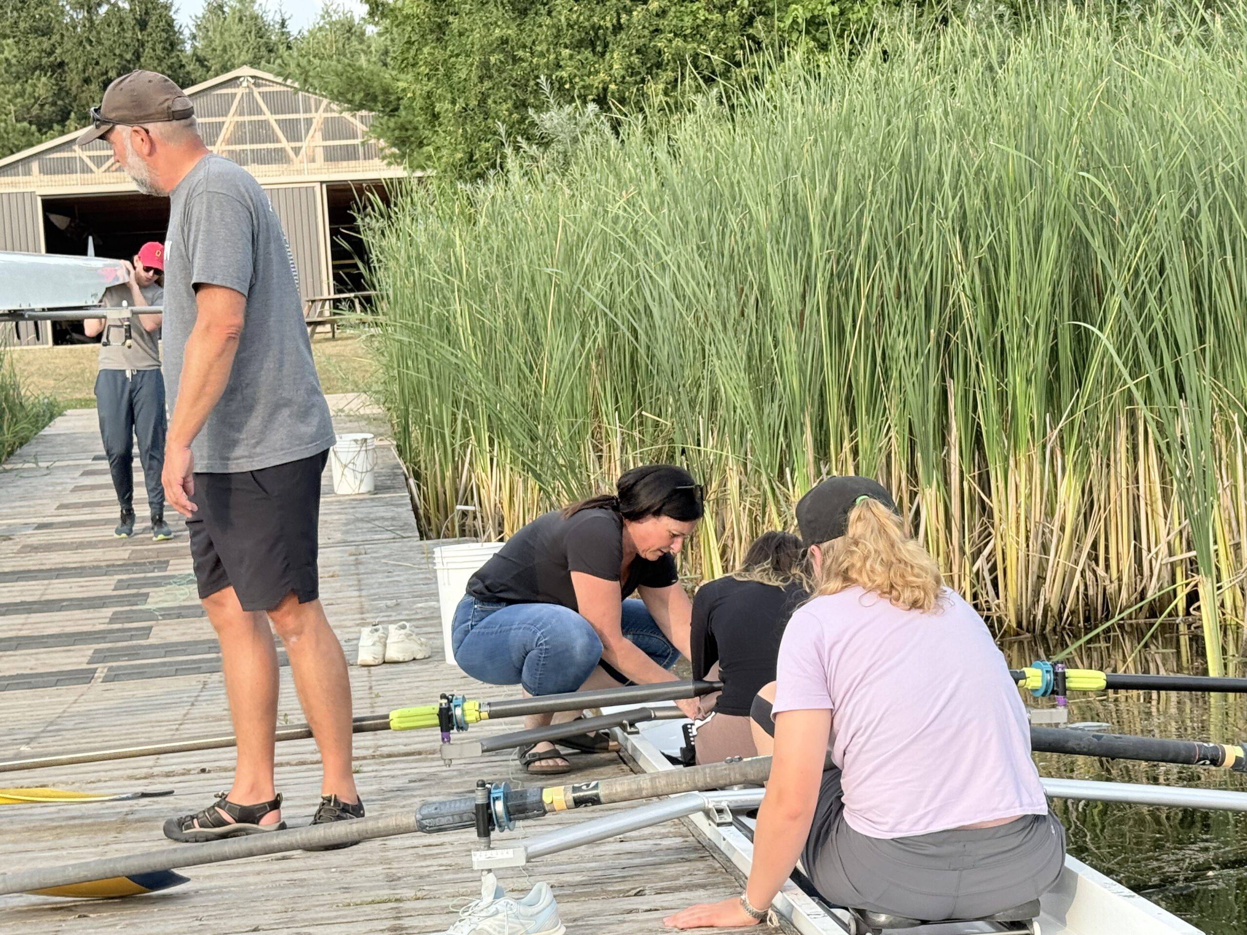 A person in a black t-shirt leans into a rowing boat to assist seated students, with others standing nearby on a wooden dock beside a lake with tall reeds.